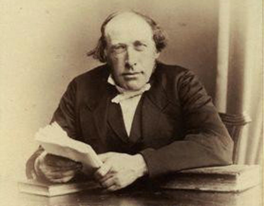 A sepia photographic portrait of the Scottish physicist and theologian Reverend John Kerr (1824–1907), discoverer of the electro-optic effect, depicted as a distinguished Victorian gentleman with a full beard, wearing a formal dark coat and white collar